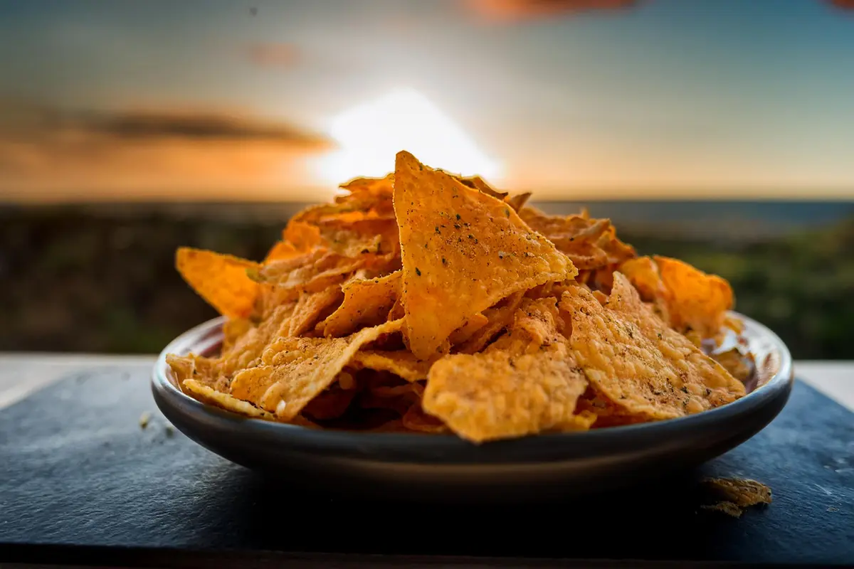 A bowl of freshly made, golden-brown Mexican chips, served with a side of salsa and guacamole, garnished with lime wedges and cilantro.